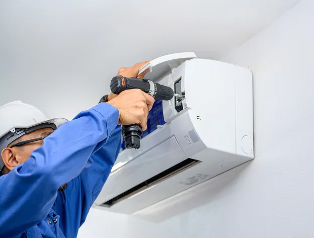 Un technicien en salopette bleue et casque blanc répare un climatiseur avec une perceuse à Buxières-d’Aillac près de Châteauroux dans l'Indre 36